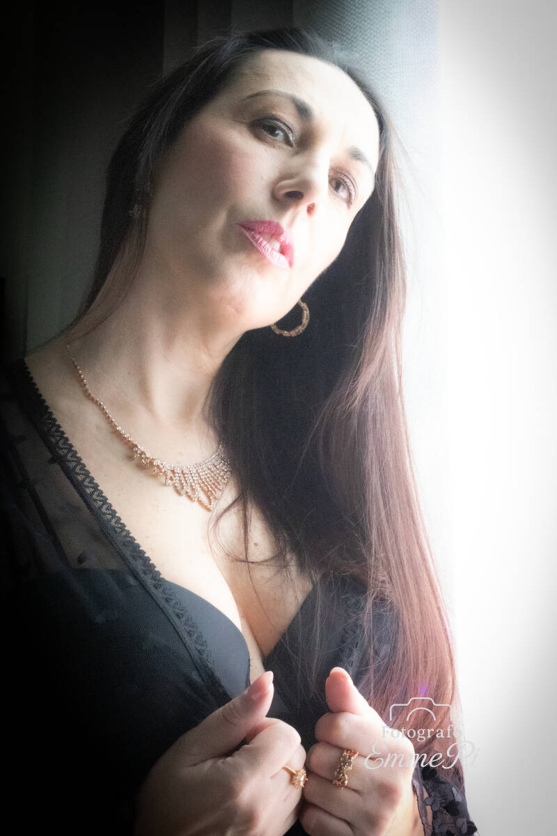 Ritratto di Margot accanto a una finestra: lunghi capelli castani, abito nero e volto illuminato dalla luce naturale, in contrasto con lo sfondo scuro.