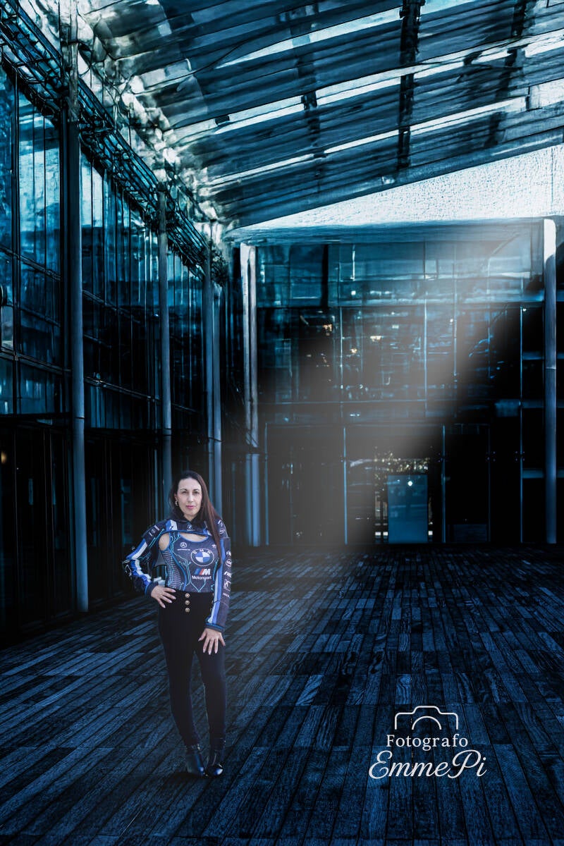 Donna con outfit sportivo blu e nero in stile motociclistico posa in un ambiente urbano futuristico illuminato da luci fredde blu, in una scena dal forte stile distopico e cinematografico.