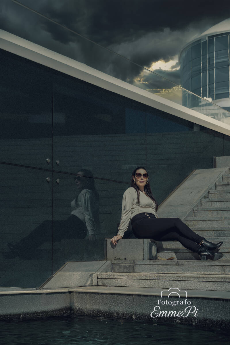 Laura seduta su una scalinata davanti a un edificio moderno di vetro, con il suo riflesso sulla parete e un cielo scuro e minaccioso sullo sfondo, in una scena dal carattere distopico e cinematografico.