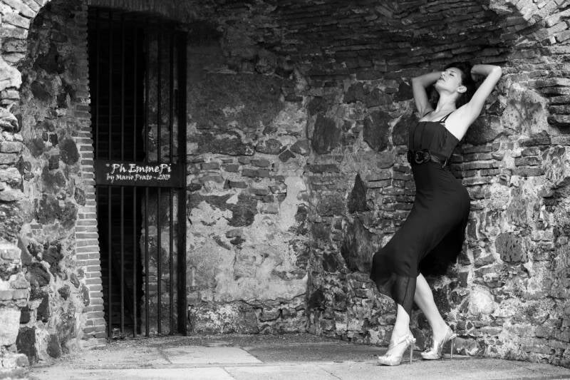 Modella in abito nero e tacchi alti posa contro un muro antico in bianco e nero, con sfondo in pietra e grata metallica.