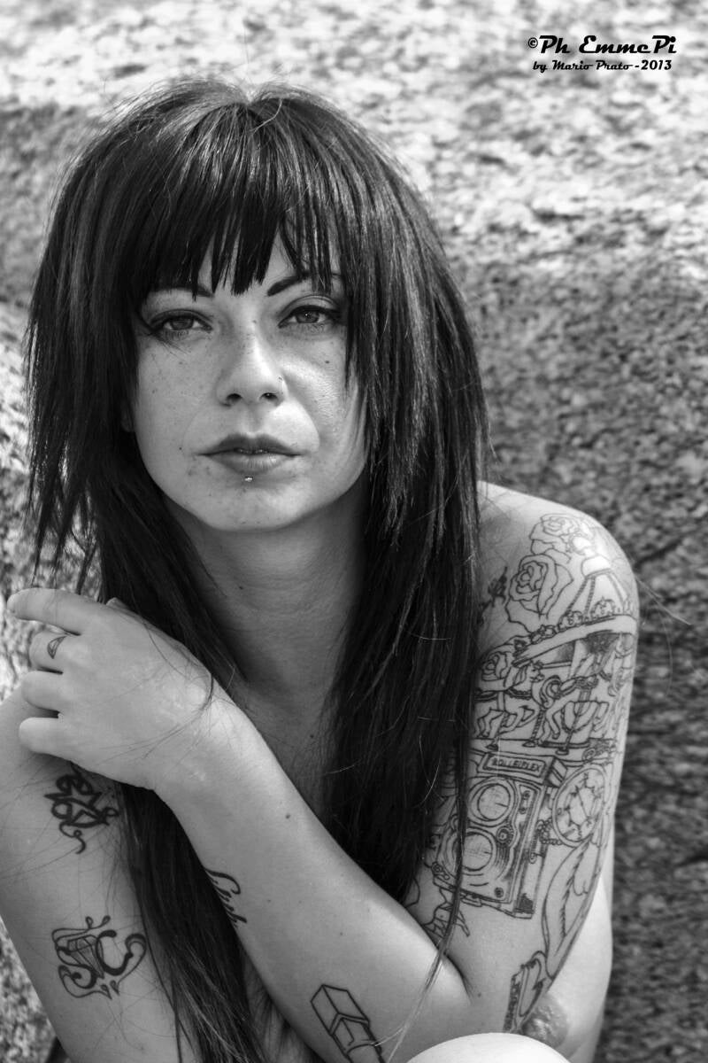 Ritratto in bianco e nero di una donna tatuata, a torso nudo con le braccia incrociate sul petto, seduta contro una roccia. Sguardo frontale e capelli lunghi con frangetta.
