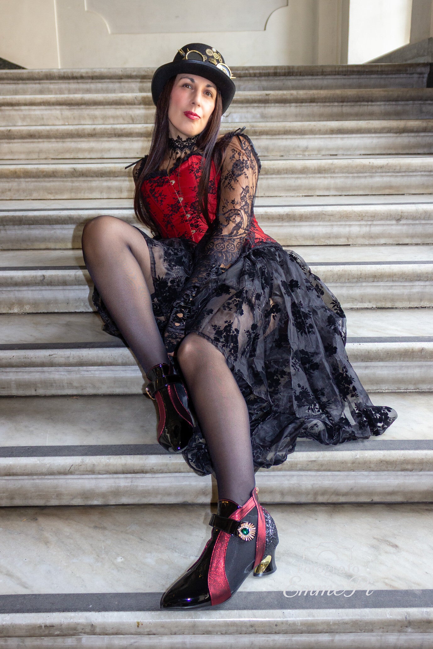 Donna in abito steampunk rosso e nero con cappello e stivaletti decorati posa seduta su scalinata in marmo