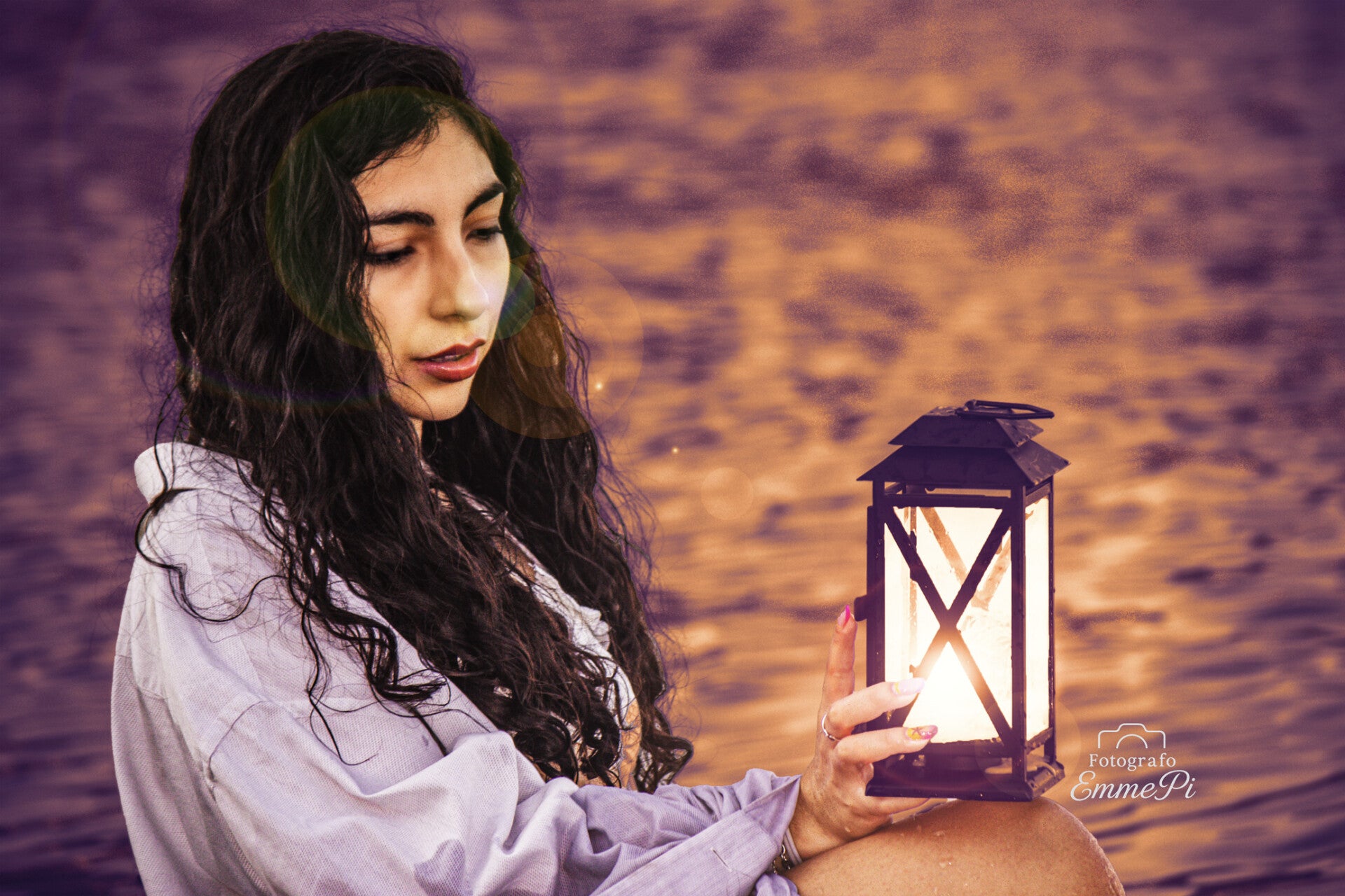 donna con capelli scuri e camicia bianca che tiene una lanterna accesa davanti al mare al tramonto