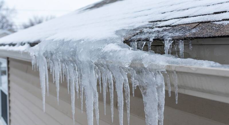 A severe winter ice dam filling a residential gutter, creating large hanging icicles and forcing trapped snowmelt to back up underneath the roof shingles.
