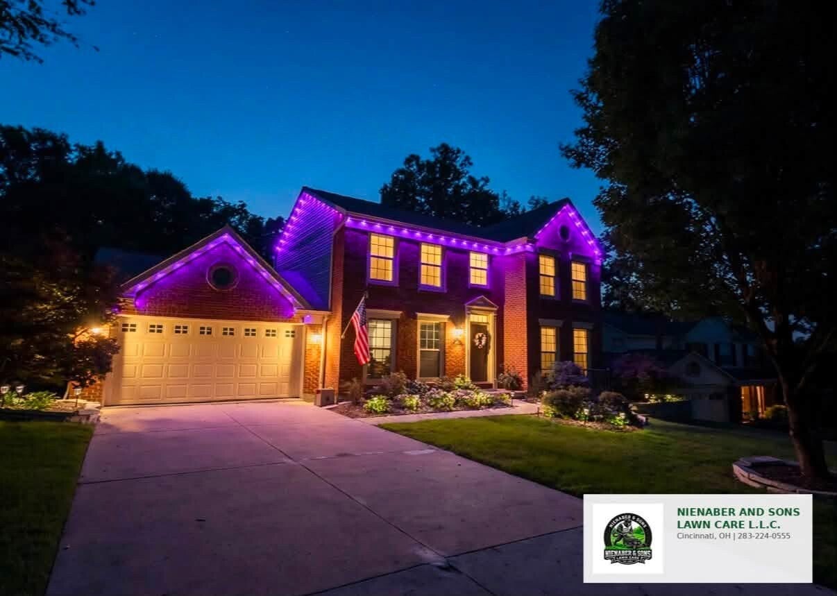 Residential brick home in Cincinnati, OH, illuminated by professional custom permanent purple LED holiday lights installed by Nienaber And Sons Lawn Care (283-224-0555), viewed at dusk with landscaping lights and a prominent company logo in the foreground