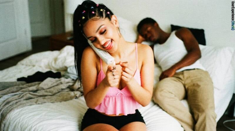 Music artist lil’ Keyah sitting on a bed, balancing a cordless phone on her shoulder and lighting a cigarillo with a man sleeping in the background.