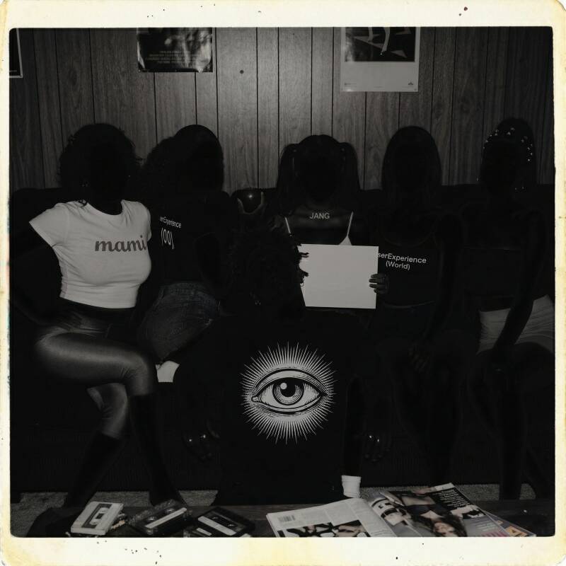 A black and white Polaroid-style photo of a group with shadowed faces in a wood-paneled room, featuring a central figure wearing a large eye graphic sweatshirt.