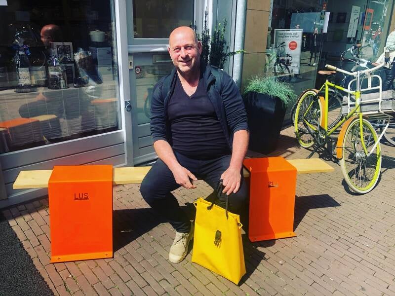 Ontwerper Leo Otten van LUS zittend op een oranje LUS bankje met de gele LUS tas