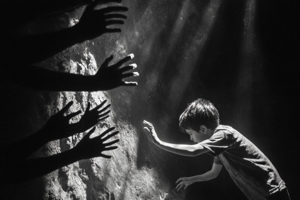 a balck and white picture of a boy trying to climb out of a dark, underground place with hands reaching out from the darkness towards him.