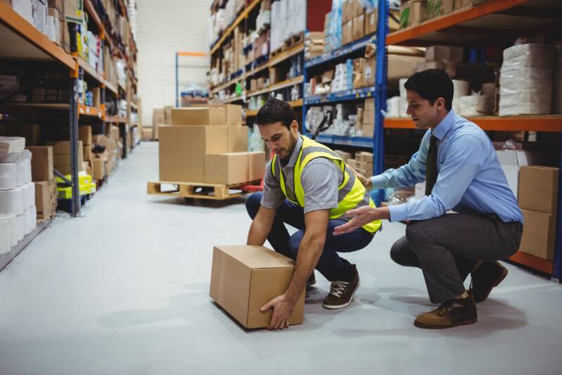 Travailleur et formateur réalisant un exercice de manutention manuelle d'un carton dans un entrepôt logistique.