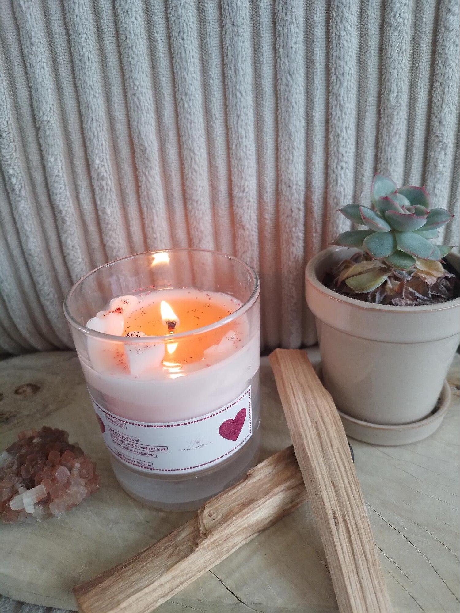 Brandende geurkaars in glazen pot met hartje, houten stokjes en roze kristal naast een vetplant op houten ondergrond.