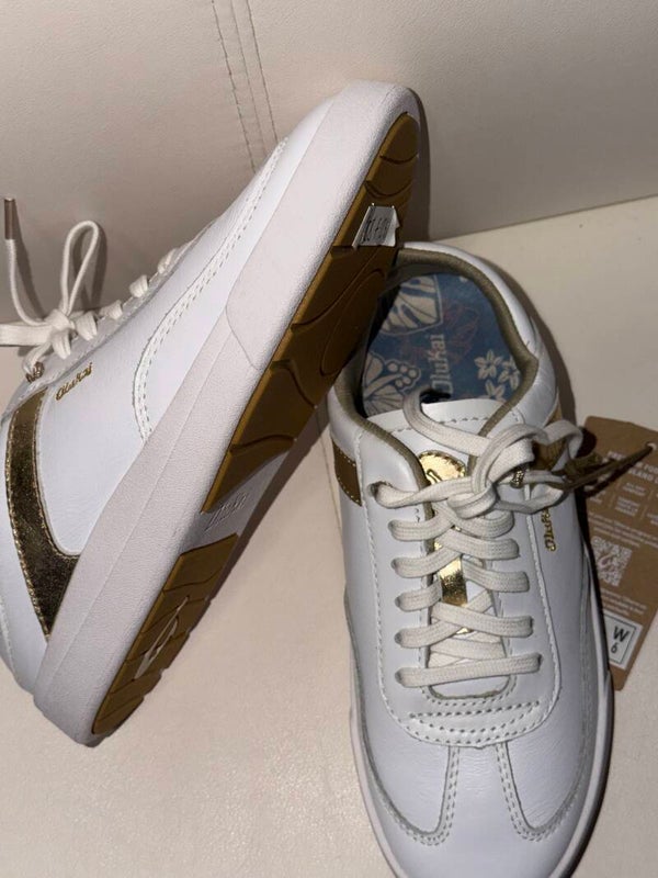 are white OluKai Women's Ha'upu sneakers featuring metallic gold detailing along