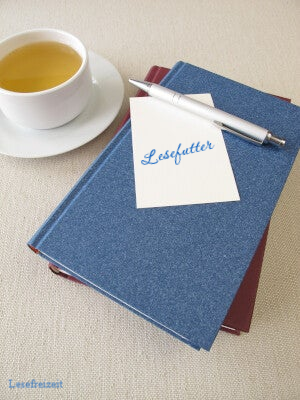 Eine Tasse Tee und ein Bücherstapel auf dem ein Notizzettel liegt, auf dem Lesefutter steht.