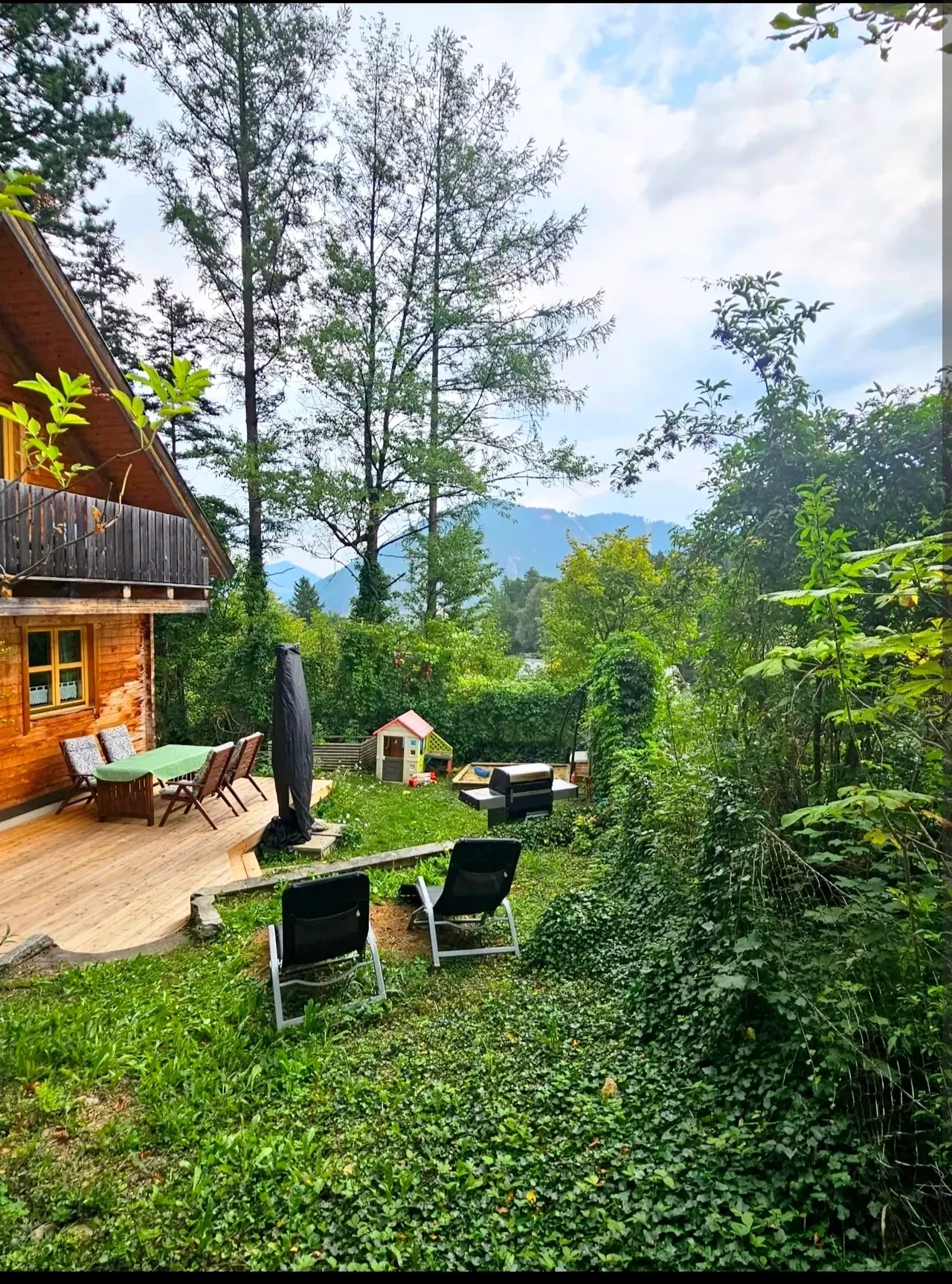 Sonniger Garten im Blockhaus Hengsttal mit Holzterrasse, gemütlichen Liegestühlen, Grillbereich und einem Kinderspielhaus im Grünen.