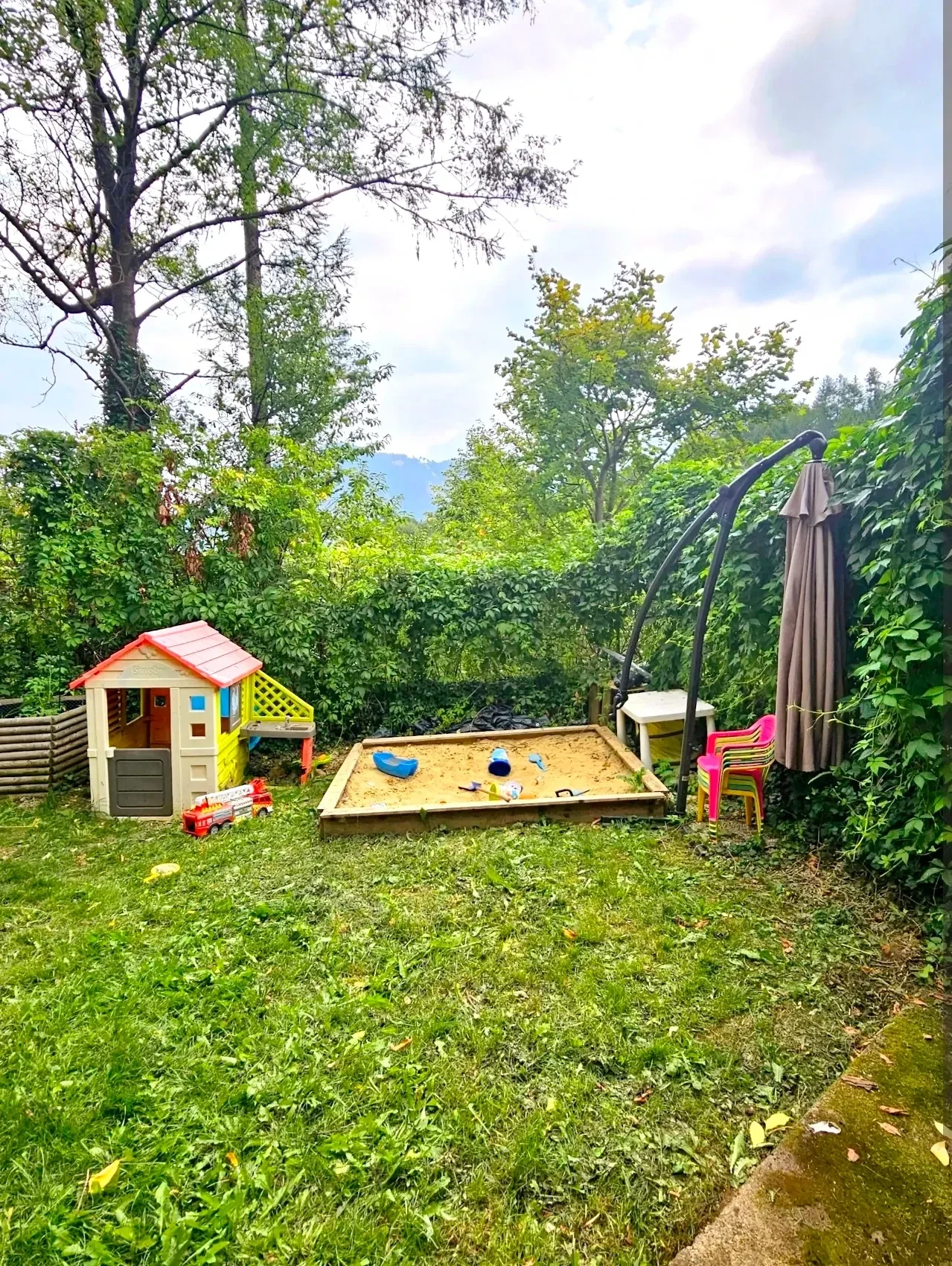 Kinderspielbereich im Garten des Blockhaus Hengsttal mit großem Sandkasten, buntem Spielhaus, Spielzeugautos und schattenspendendem Sonnenschirm