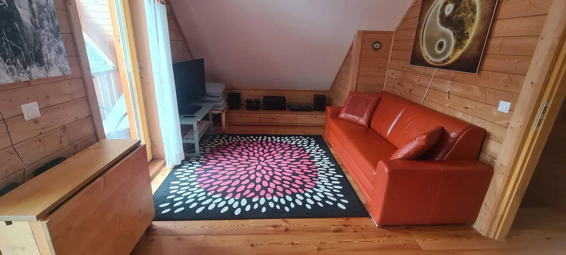 Die andere Seite des Schlafzimmers: Gemütliche TV-Lounge im Obergeschoss des Blockhaus Hengsttal mit rotem Ledersofa, modernem Teppich u mit einem Tisch der gerne als Arbeitsplatz verwendet wird.