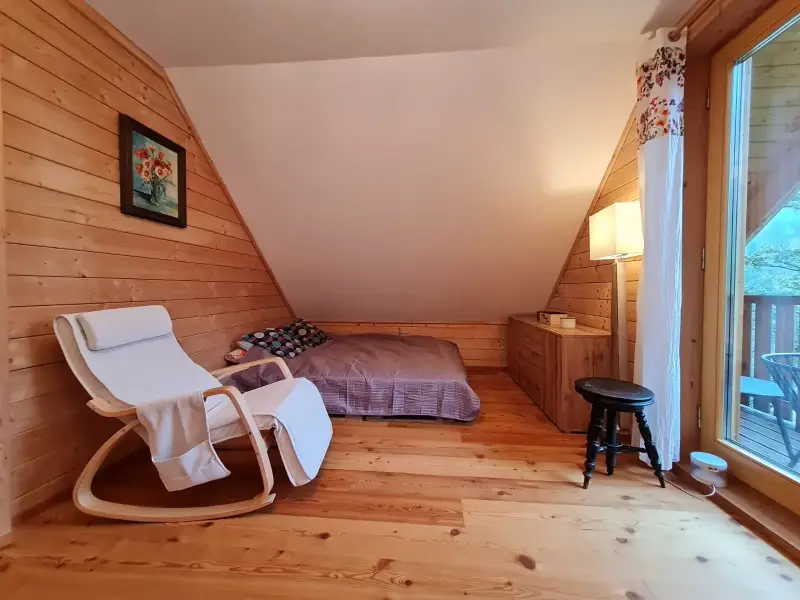 Gemütliches Schlafzimmer im Obergeschoss des Blockhauses mit Dachschräge, Lesestuhl und Zugang zum Balkon mit Aussicht.