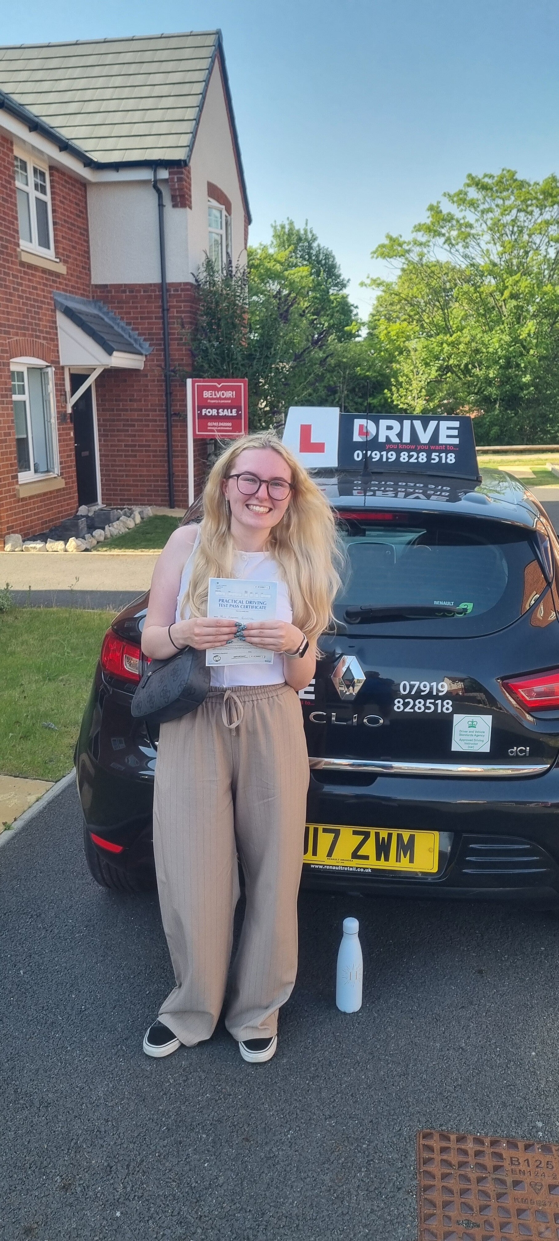 Happy driving school student holding a practical driving test pass certificate outside the tuition car.