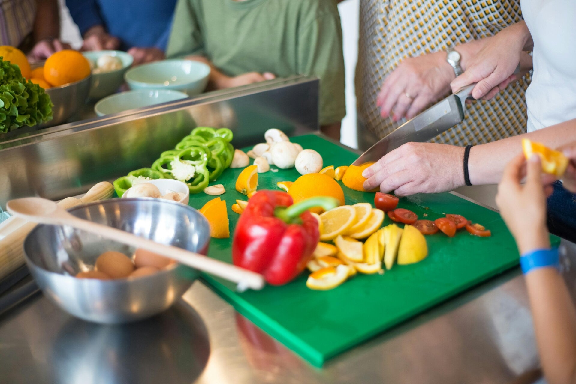 Hände schneiden frisches Gemüse wie Paprika und Zitronen auf einem grünen Schneidebrett bei einem Kochkurs.