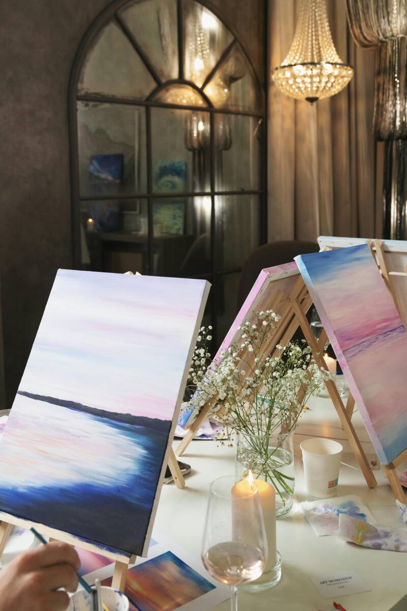 Mehrere Staffeleien mit bunten Leinwänden, Pinseln und Weingläsern stehen für ein Sip und Paint Event bereit.