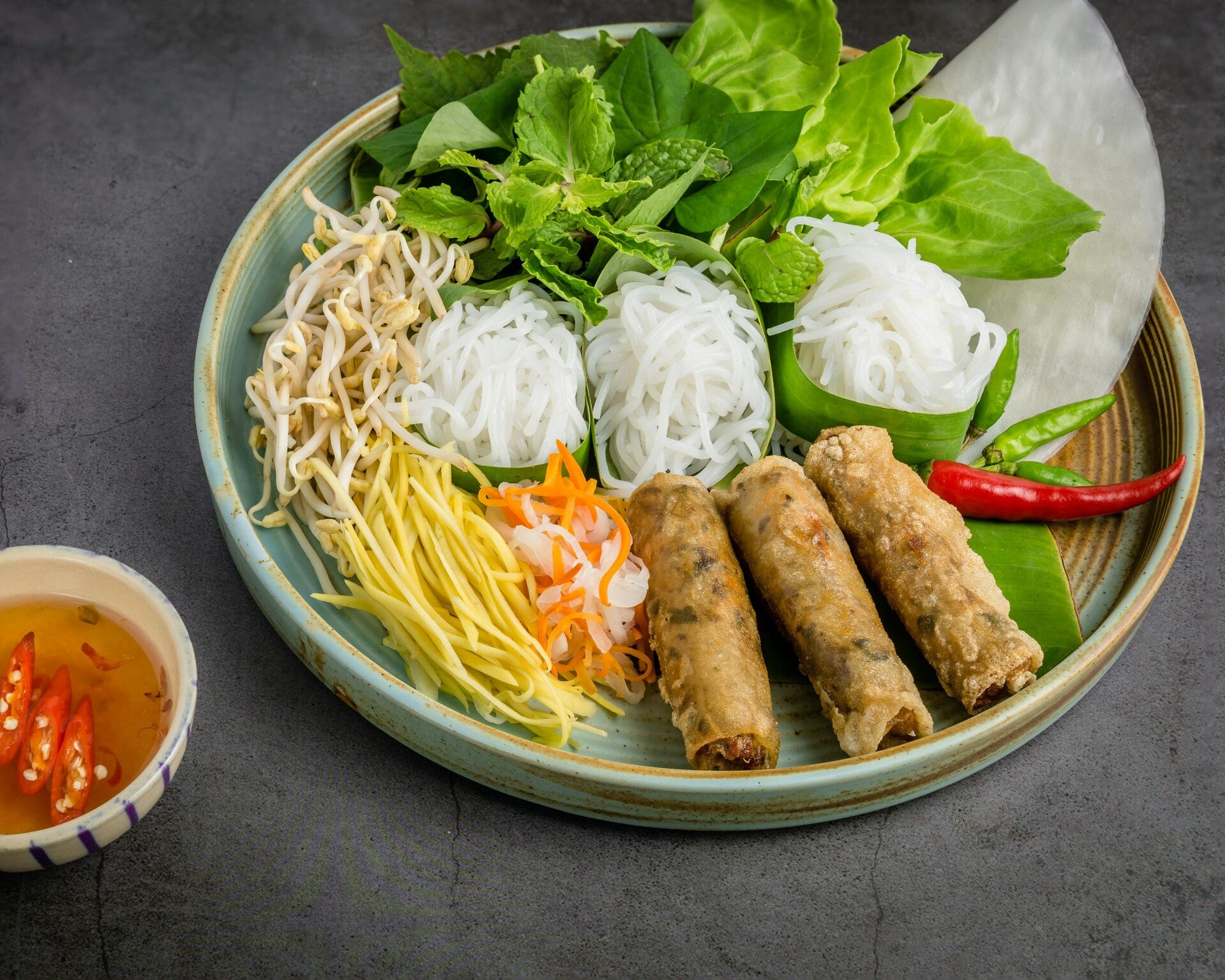 Ein Teller mit vietnamesischen Frühlingsrollen, frischen Kräutern, Sprossen, Nudeln und Reispapier.