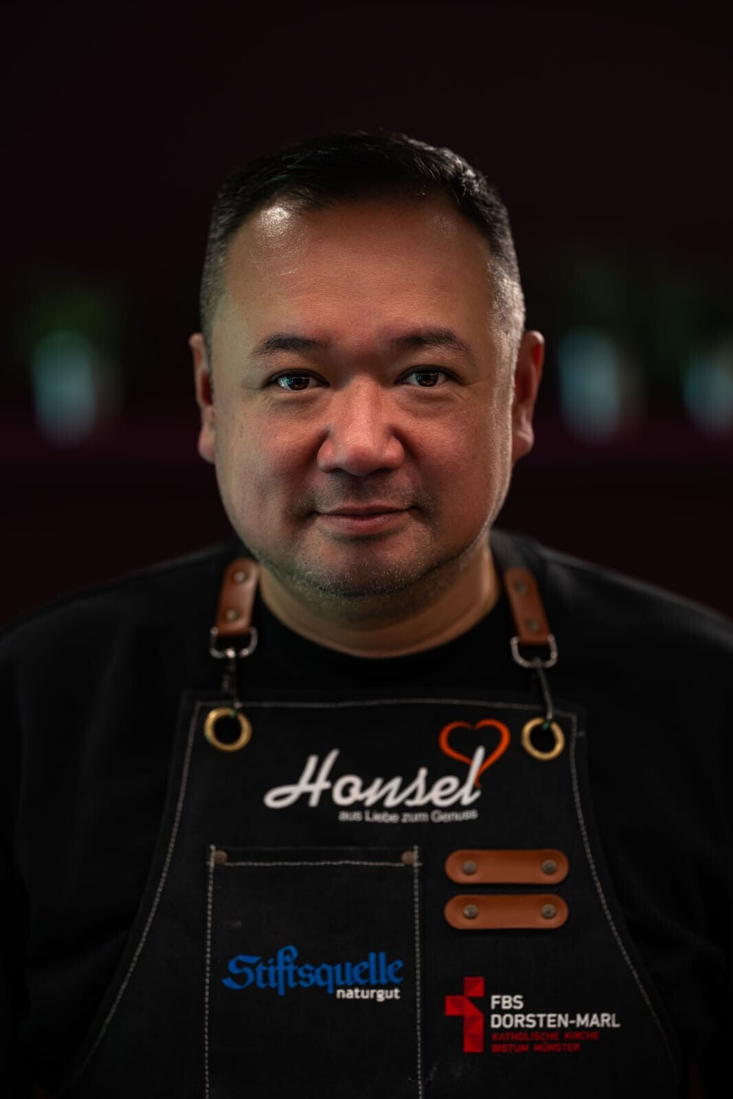 Minh Lam (MasterChef 4. Platz & Küchenschlacht 2. Platz) in Honsel-Schürze vor Flammenhintergrund.