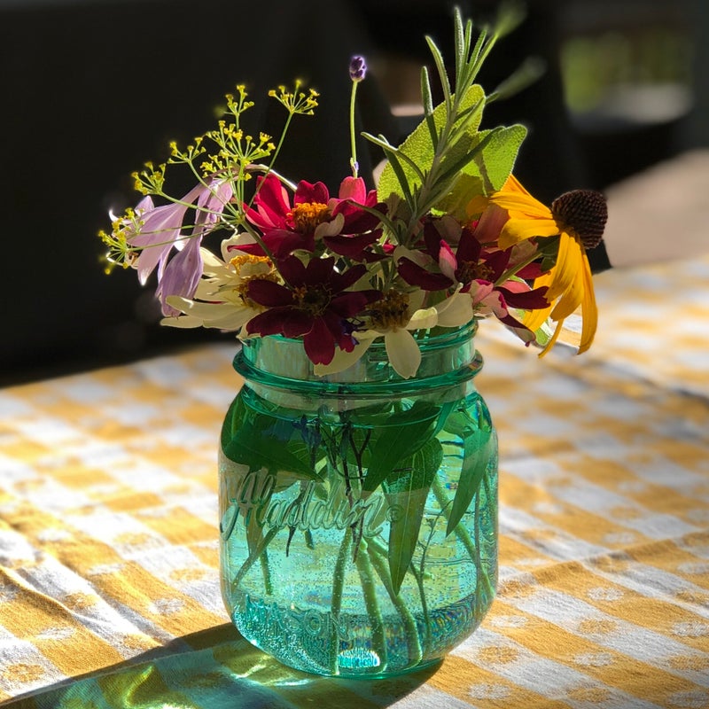Short blue jar filled with a floral arrangement featuring yellow dill blooms, small deep red and white zinnia, black eyed Susan, lavender, rosemary and mint on top of a gold and white striped and floral vintage tablecloth. The sun illuminates it from behi