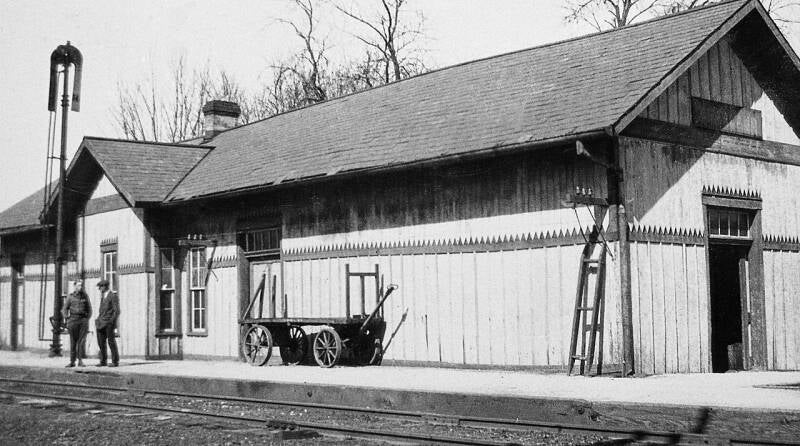 a stayion building, freight wagon and two figures standing trackside