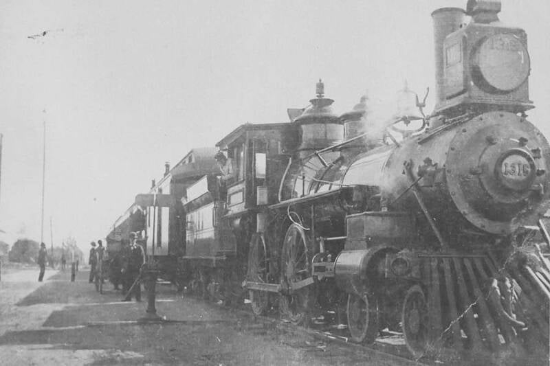black and white photo of a 4-4-0 steam locomotive with tende pulling passenger train with several people standing trackside