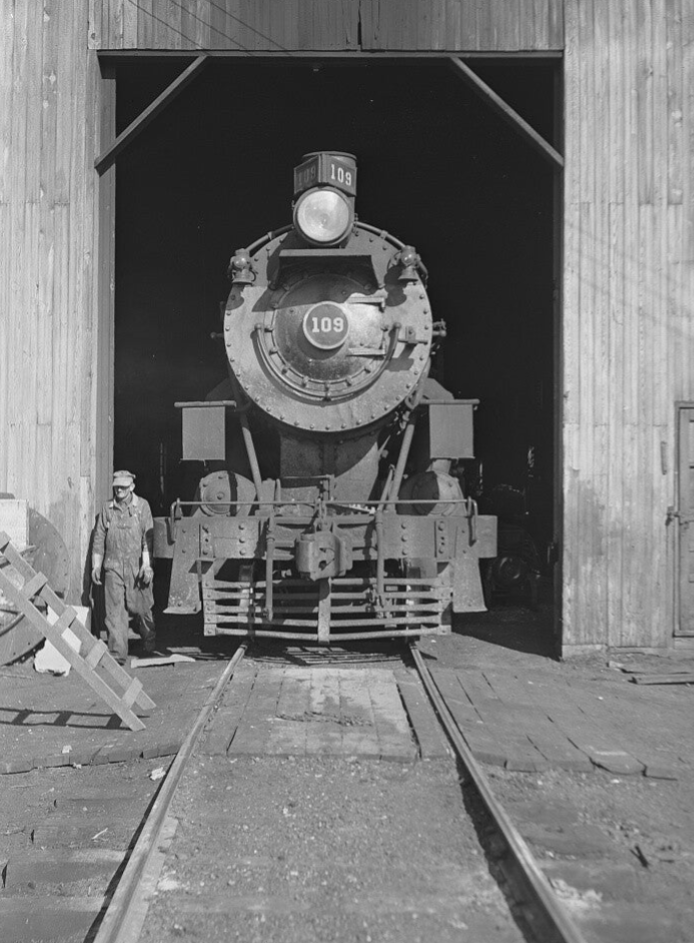 a crewman walks trackside as a large steam locomotive sits in an enginehouse