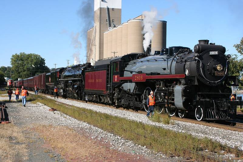 two steam locomotives pulling a passenger train by some grain elevators