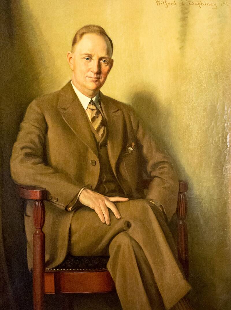 A well-dressed man in a brown suit sitting in a chair.