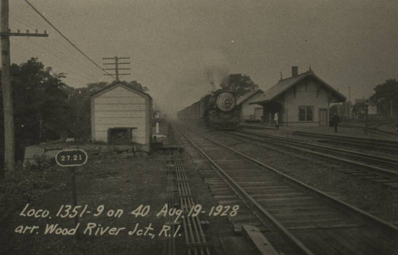 A steam locomotive pulls a train next to a small train station. Several tracks are visible.