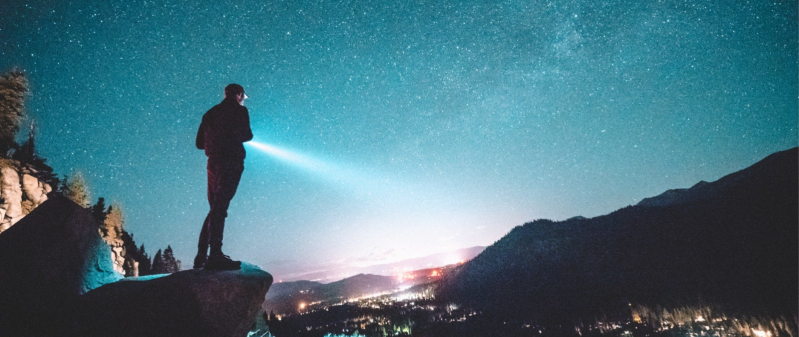 A person standing on a rocky cliff at night, holding a flashlight pointed toward the horizon. The sky above is filled with stars, and a distant city glows below in the valley, surrounded by mountains.