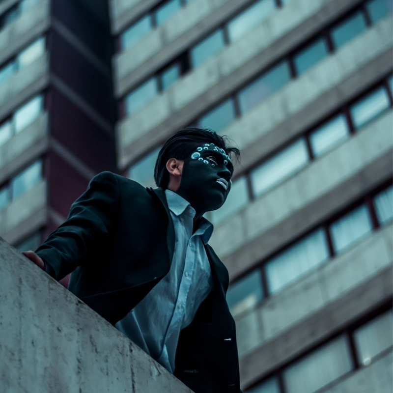 Person in business attire with black face paint and googly eyes, standing near a concrete wall, looking out pensively. A tall building with numerous windows looms in the background