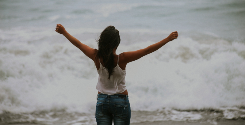 Person standing with arms raised, facing the ocean waves