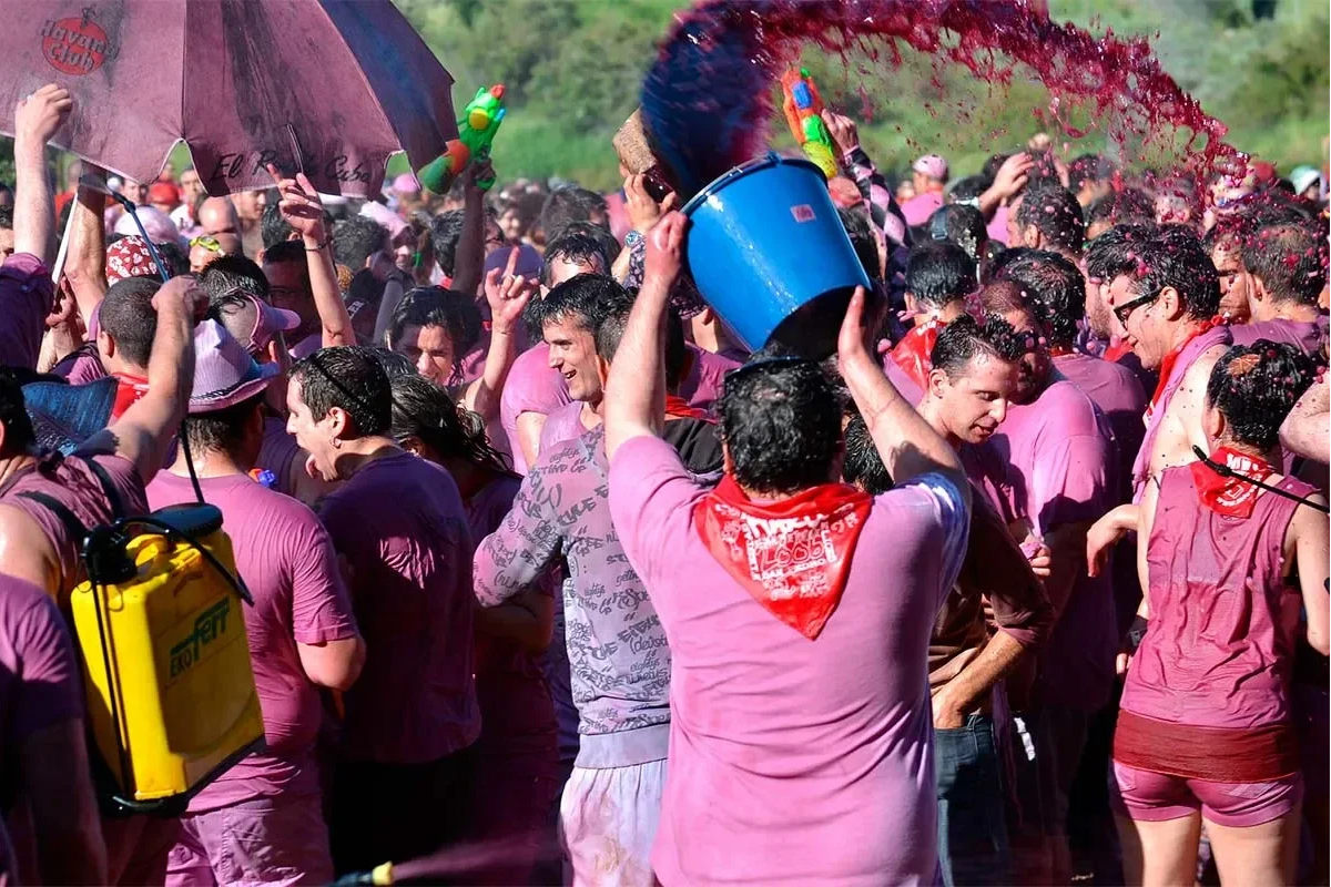 Batalla del vino con muchas personas tirandose cubos de vino, las camiseta blancas son rosas y todos llevan un pañuelo rojo en el cuello, se lo estan pasando fenomenal