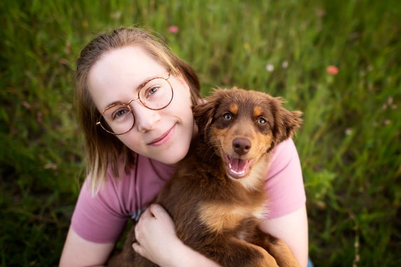 Een vrouw met een bril en zachtroze t-shirt knuffelt een vrolijke red tri Australische Herder puppy in een groen veld.