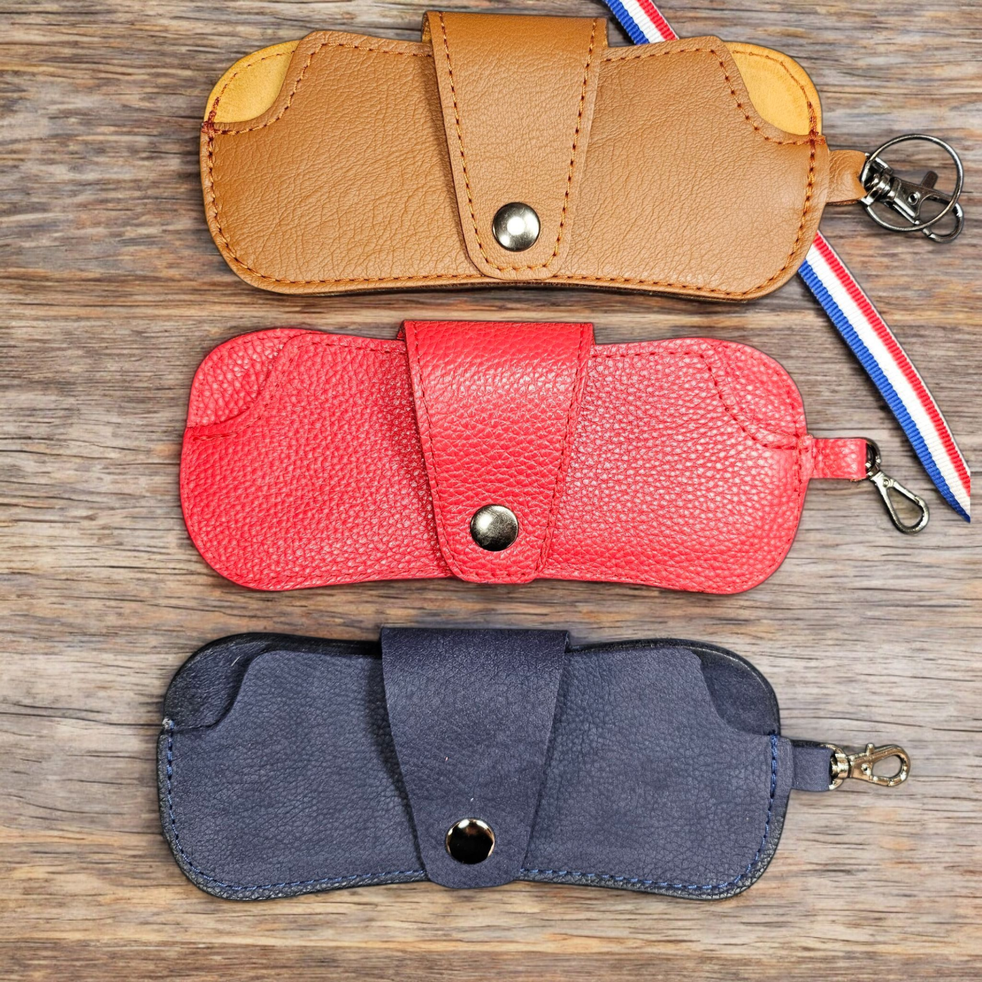 Trois porte lunettes en cuir. Un cognac, un rouge et un bleu avec un liseret bleu blanc rouge. Posés sur une table en bois