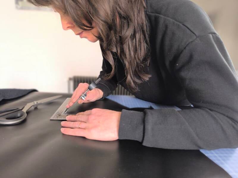 Artisane du cuir travaillant sur un cuir noir, outil à la main. Une paire de ciseaux trône sur la table de travail