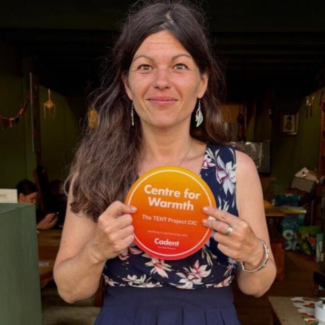 A dark haired lady called Victoria standing in the cafe doorway holding a round Candent Centre For Warmth Award Plaque.