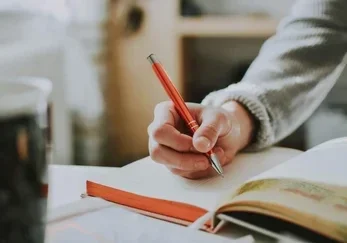 A close-up of a hand holding a red pen while writing in a notebook, depicting Act III: The Writing and the creation of a new life chapter.