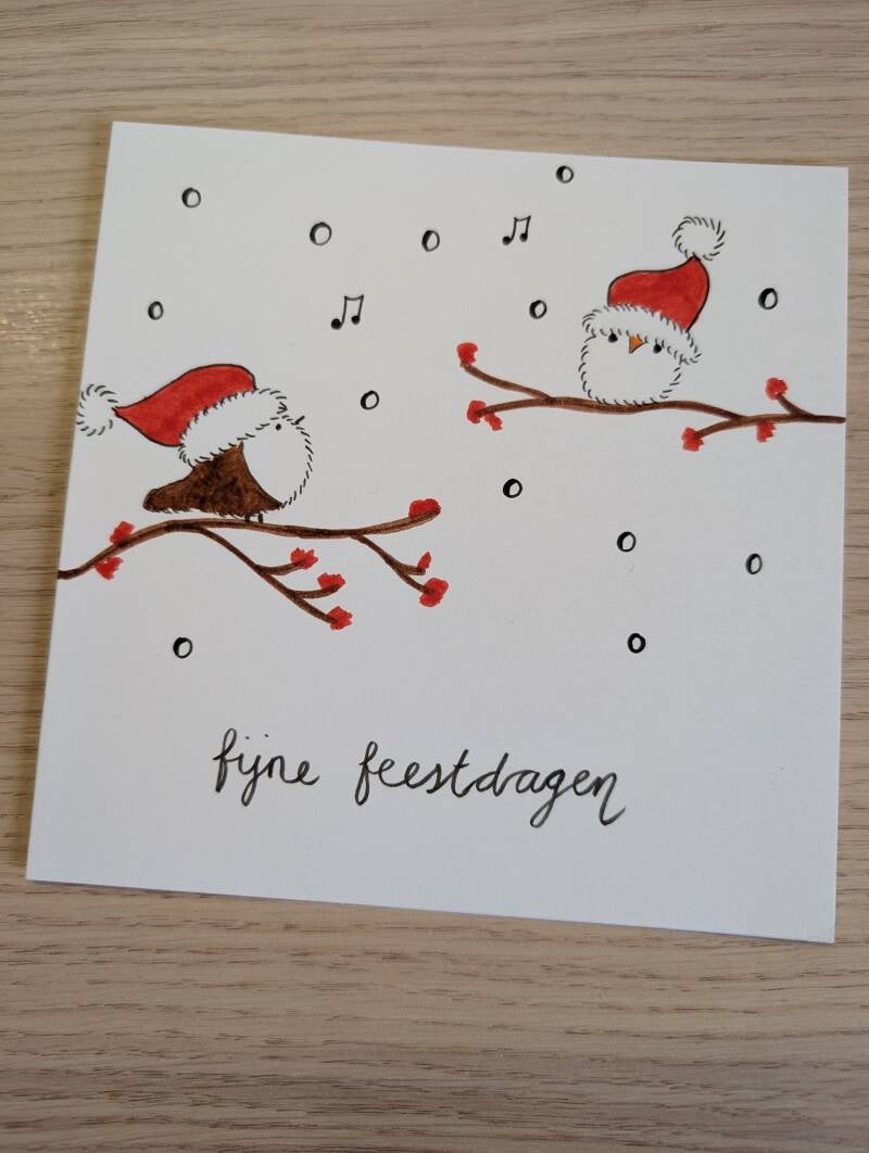 witte kaart met twee vogeltjes op een tak met allebei een kerstmuts op, terwijl het sneeuwt. Met de tekst Fijne feestdagen eronder.