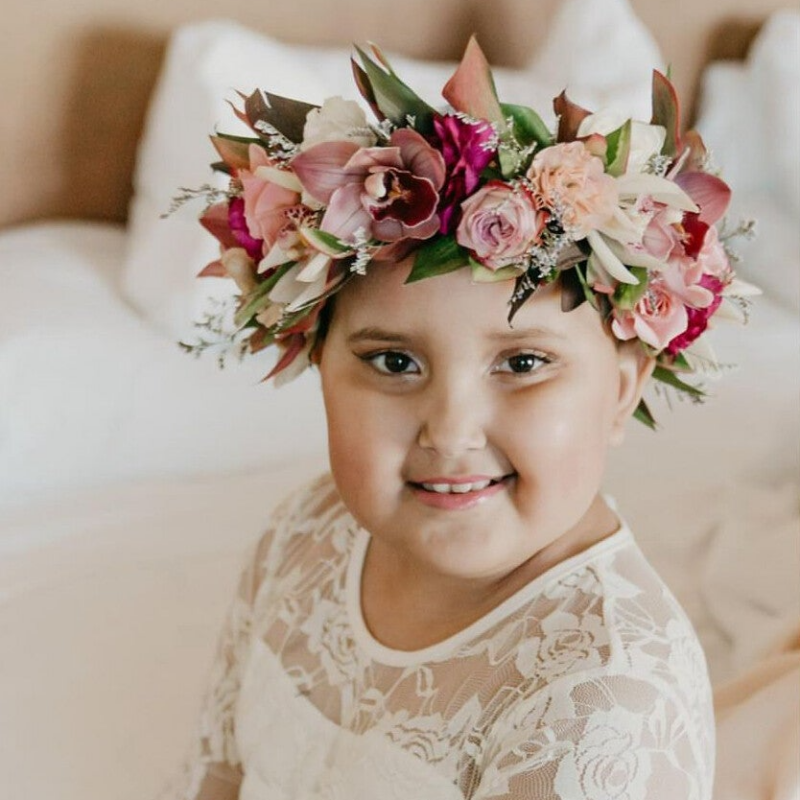 A photo of yooung girl in white floral dress  wiith a flower crown on. She is siting on a white fluffy chair with her arms crossed. She has bald head and is smiling.
