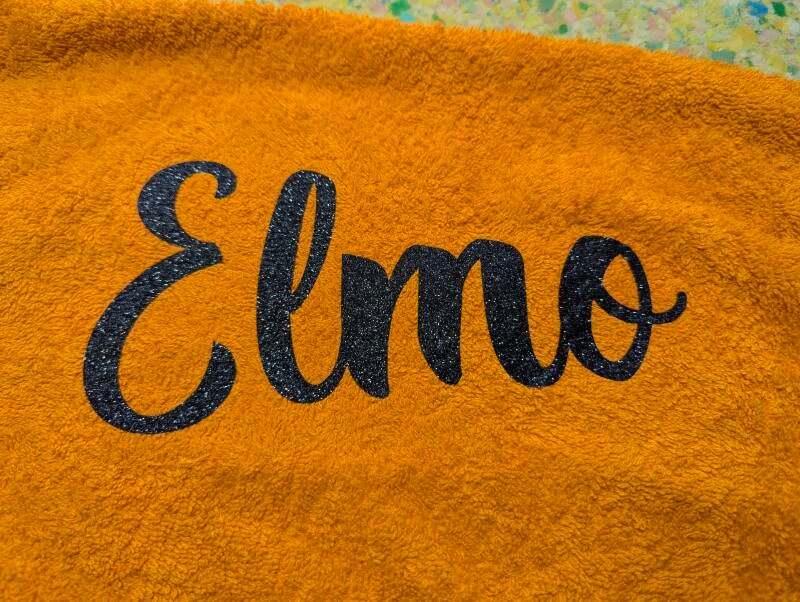 Personalisierte Schrift Elmo in Glitzer Schwarz für ein Hundehandtuch in orange.