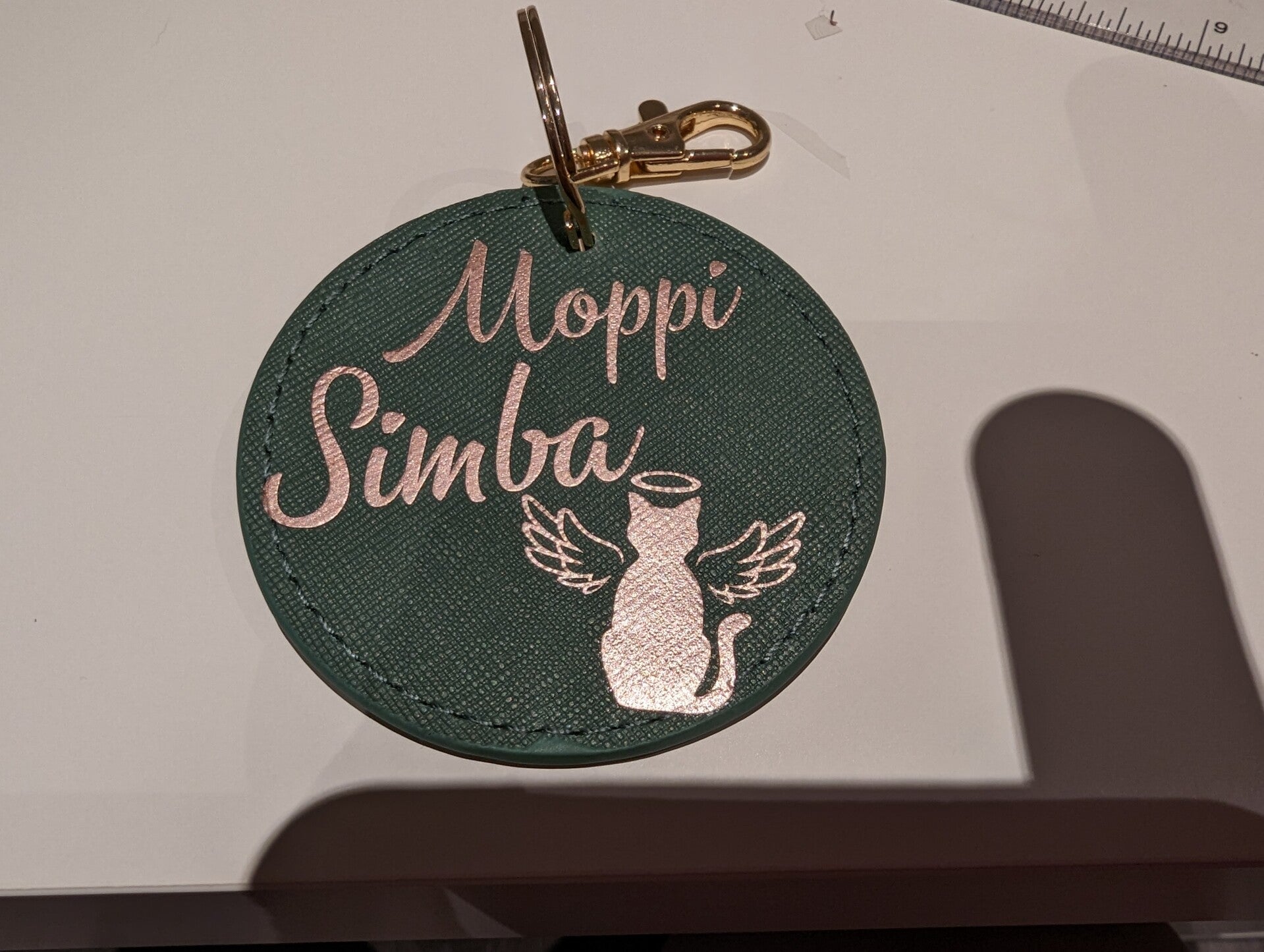 Personalisierter Schlüsselanhänger in rund und grün mit rosegoldener Aufschrift Moppi und Simba und einer Katze mit Engelsflügel.