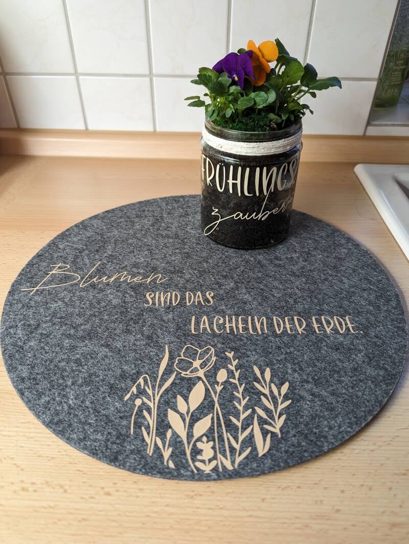 Runder mittelgrauer Filzuntersetzer mit Blumen in beige und der Aufschrift Blumen sind das Lächeln der Erde in gleicher Farbe. Dekoriert mit einem Einmachglas mit Blumen.