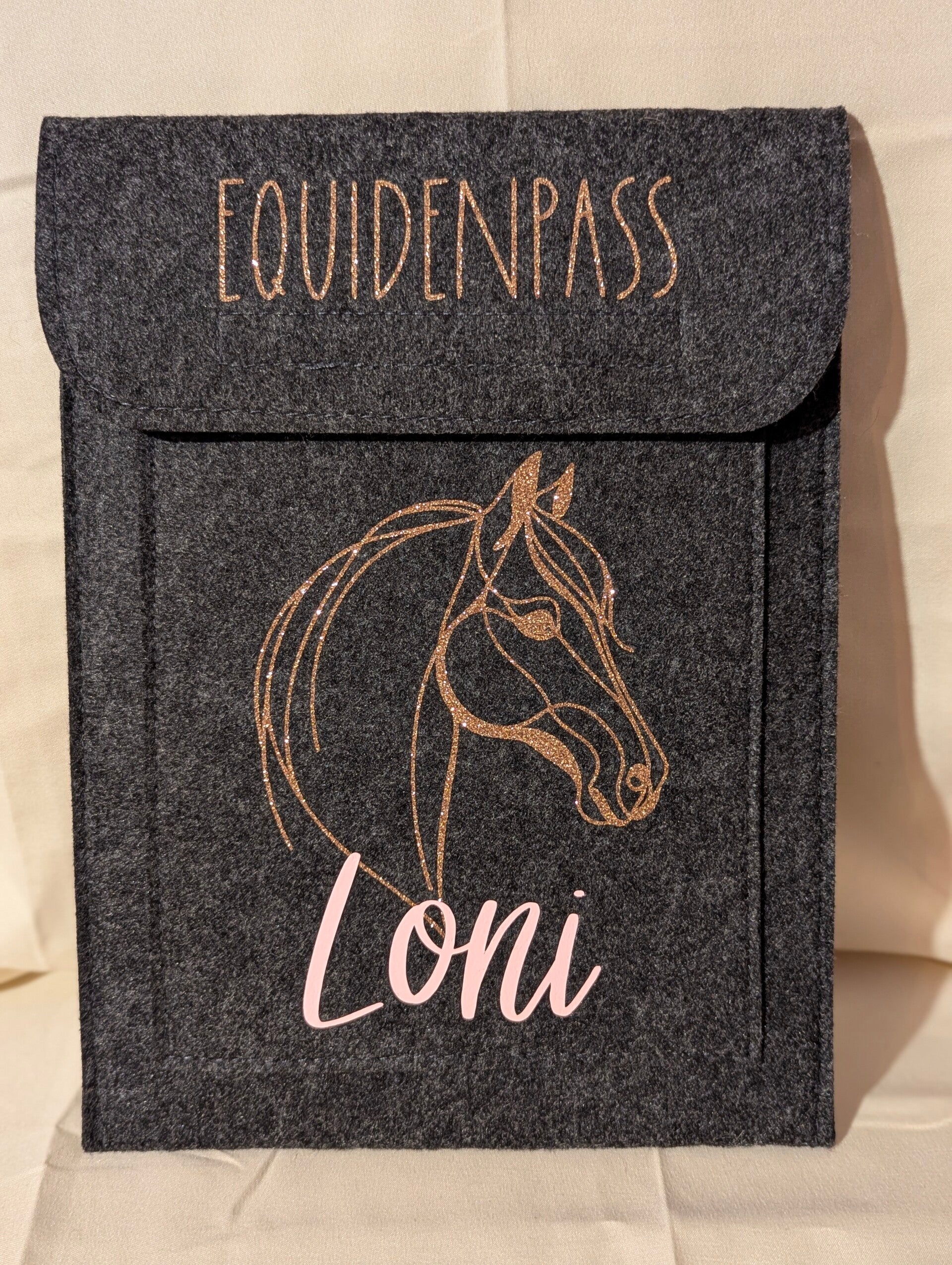 Filz Equidenpasshülle in dunkelgrau mit Aufschrift Equidenpass in gold Glitzer, einem Pferdekopf in Gold Glitzer und der Aufschrift Loni in rosa.