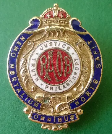 Oval gold-toned enamel badge with a crown. Centre features a red circle with gold letters spelling "RAOB". Surrounding the centre is a white ring with the words "JUSTICE TRUTH PHILANTHROPY" in gold lettering. Outer dark blue enamel border.