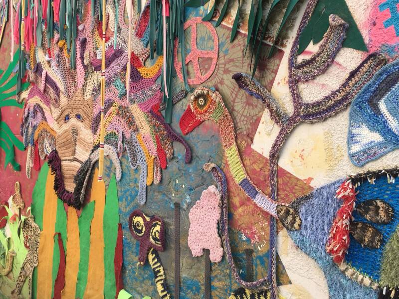 De afbeelding toont een kleurrijke muurkunstinstallatie gemaakt van textiel en haakwerk, vaak aangeduid als 'yarn bombing' of straatkunst. 